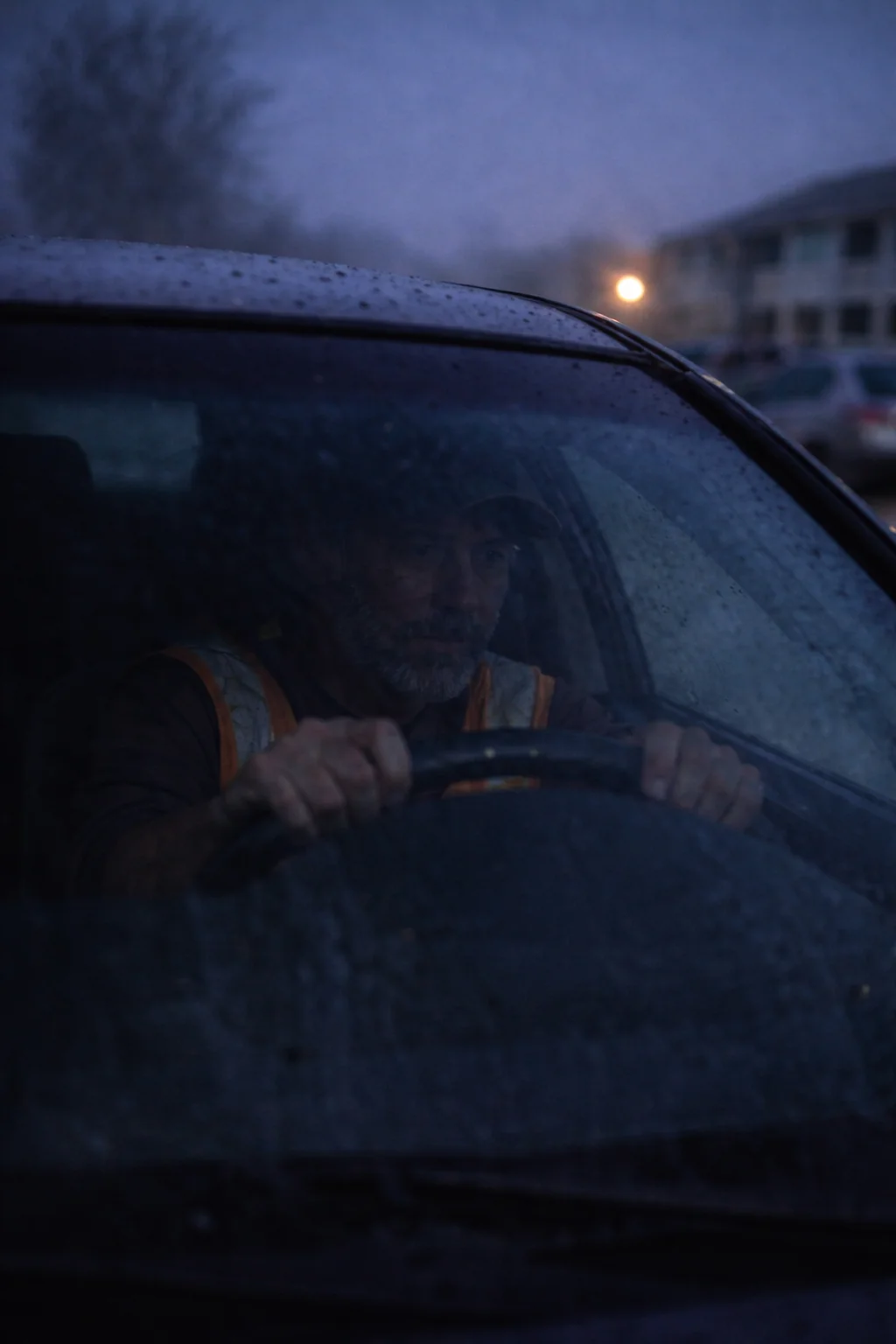 Worker gripping a steering wheel before dawn, looking through a rain-spotted windshield